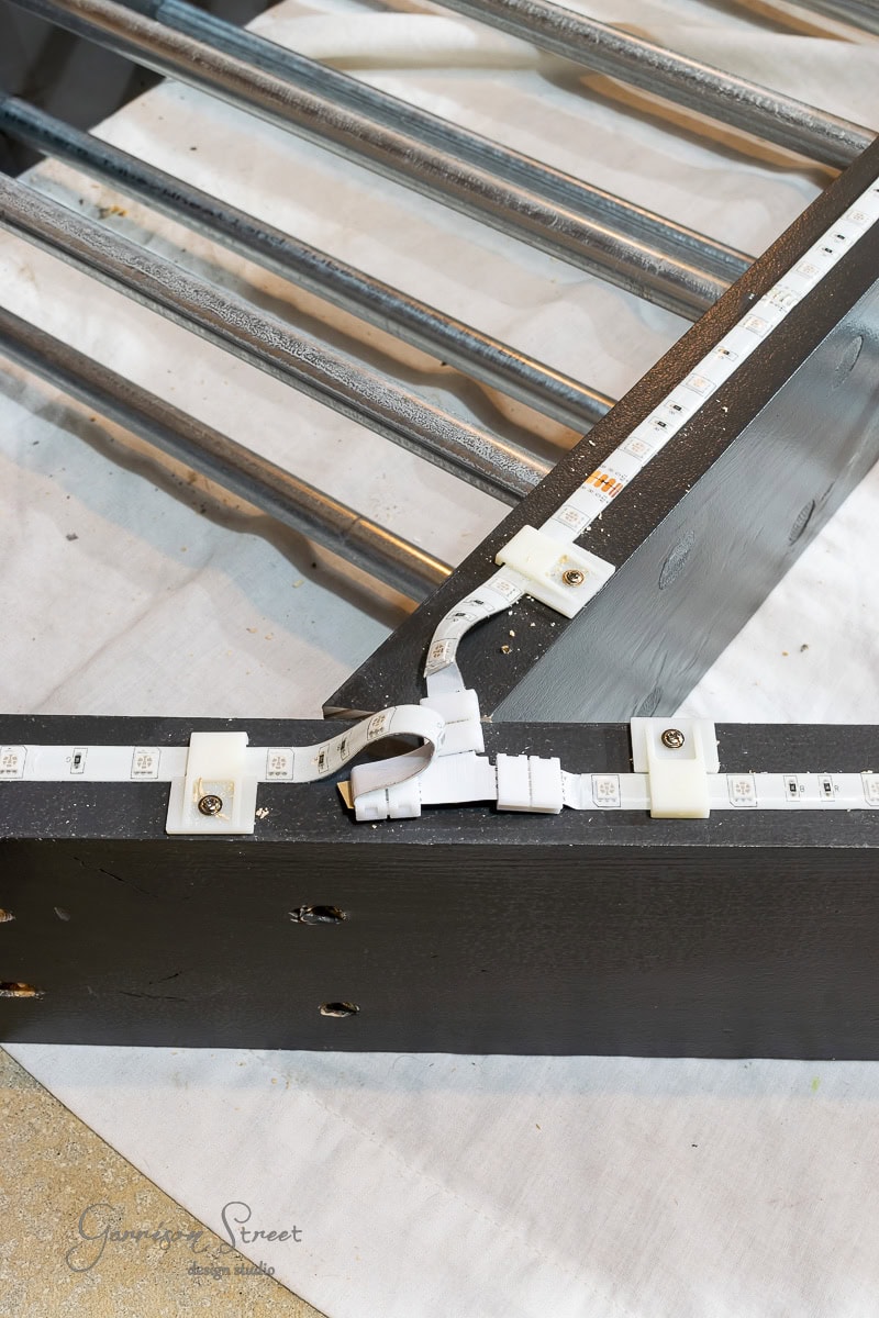 Close-up of a black wooden frame with metal slats, reminiscent of building a DIY wine rack. Attached is a strip of LED lights with connectors. The LEDs have a white strip, fixed with small white clips, all elegantly set on a white sheet.
