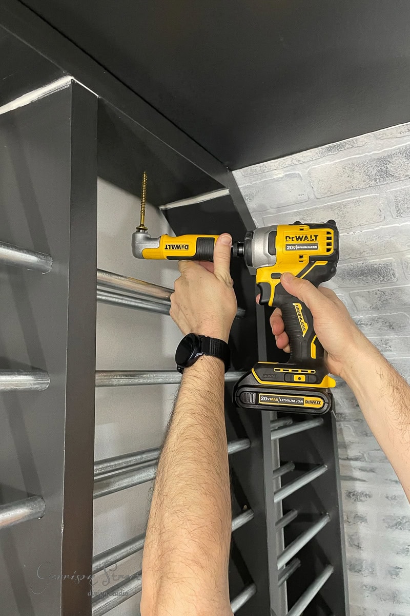A person uses a yellow and black DeWalt power drill to assemble a gray metal shelf, akin to building a DIY wine rack. The drill is positioned near the screw being tightened against the light gray brick-textured wall in the background.