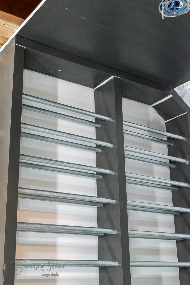 A close-up of a metal shelving unit with horizontal rods, reminiscent of steps in how to build a DIY wine rack. The shelves have a dark gray frame and are positioned vertically, set against a light-colored background.