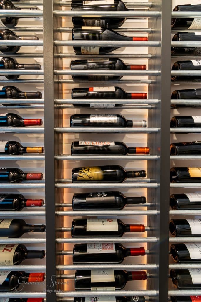 About A metallic vertical wine rack filled with various bottles, showcasing colorful labels and caps, stands elegantly against the wall. This decorative storage solution is perfect for your collection and might inspire you to learn how to build a DIY wine rack of your own.