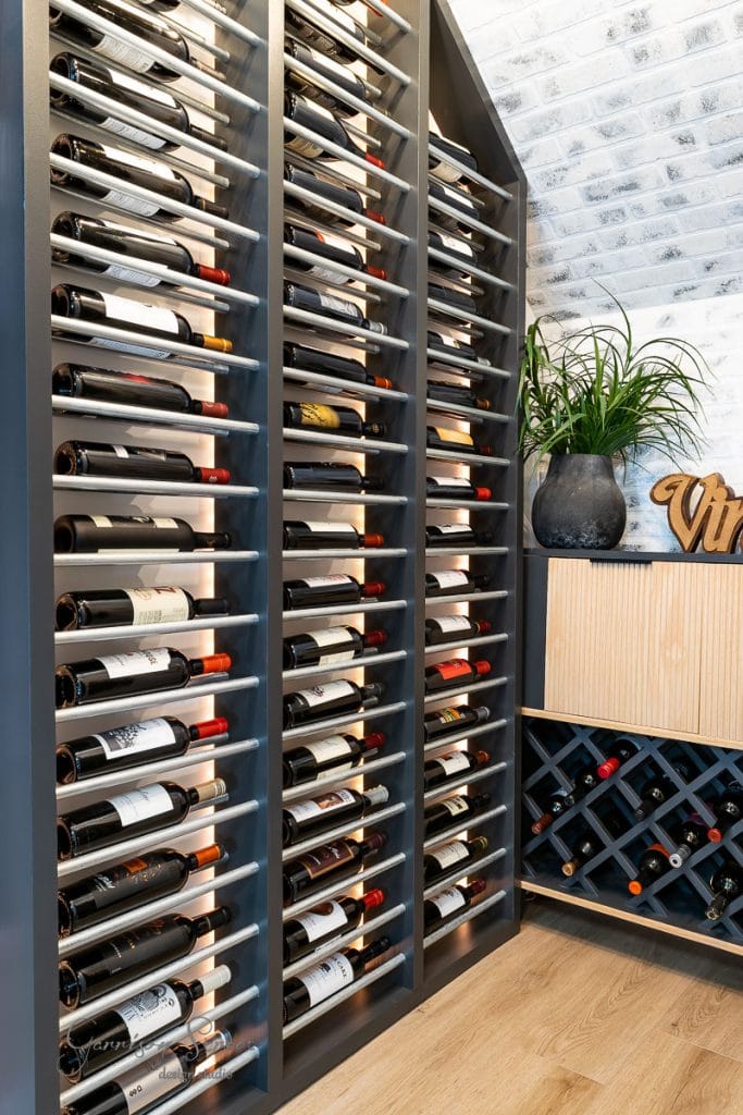 A modern wine cellar showcases rows of wine bottles stored horizontally on dark shelves, reminiscent of the elegance found in guides on how to build a DIY wine rack. A small wooden cabinet to the side holds more bottles and a black pot with green plants on top. The floor is wooden, and the white wall features a brick pattern.