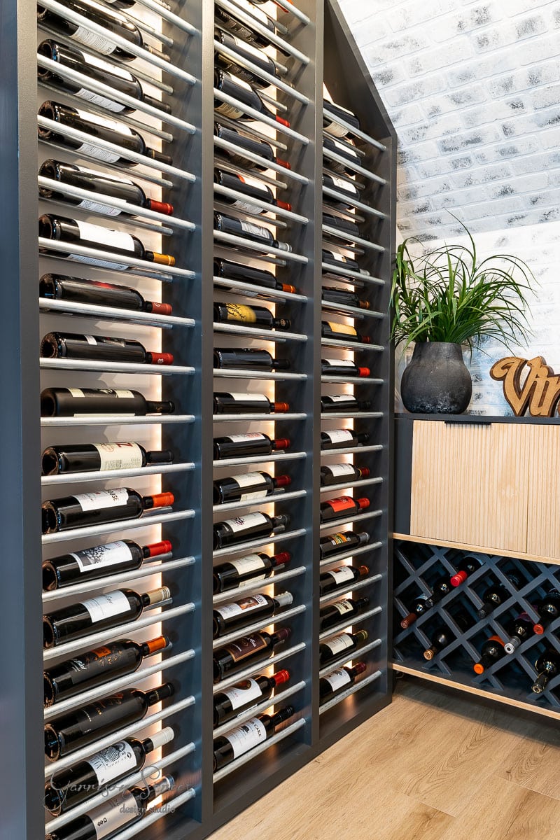 A modern wine cellar showcases rows of wine bottles stored horizontally on dark shelves, reminiscent of the elegance found in guides on how to build a DIY wine rack. A small wooden cabinet to the side holds more bottles and a black pot with green plants on top. The floor is wooden, and the white wall features a brick pattern.