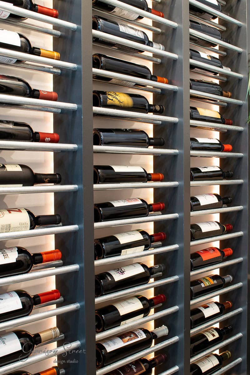 Wall-mounted wine racks filled with bottles, each resting horizontally, offer DIY inspiration. The shelves are made of metal rods, showcasing a variety of wines with different colored labels and caps. This neat and organized display exemplifies how to build a stylish wine rack at home.