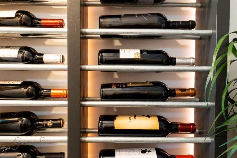A wine rack filled with horizontally arranged bottles showcases various labels and colors. With metal bars partially illuminated, it hints at an elegant DIY design. A green plant graces the right side, adding a touch of nature to inspire your own DIY wine rack project.