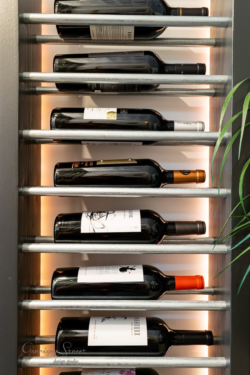 Learn how to build a DIY wine rack that elegantly holds nine horizontal bottles with assorted labels, softly illuminated by backlighting. A touch of nature is added by a green plant peeking from the right, and the rack is stylishly mounted on a dark wall.