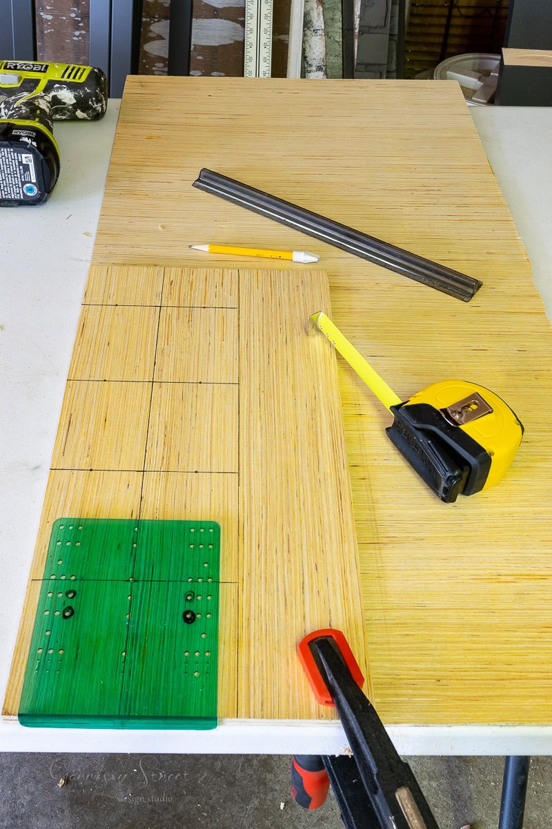 A woodworking setup for crafting a DIY wine rack includes a piece of wood with grid lines, a yellow tape measure, a pencil, a ruler, and a green measuring tool secured by a clamp on the white table. A power tool looms in the background.