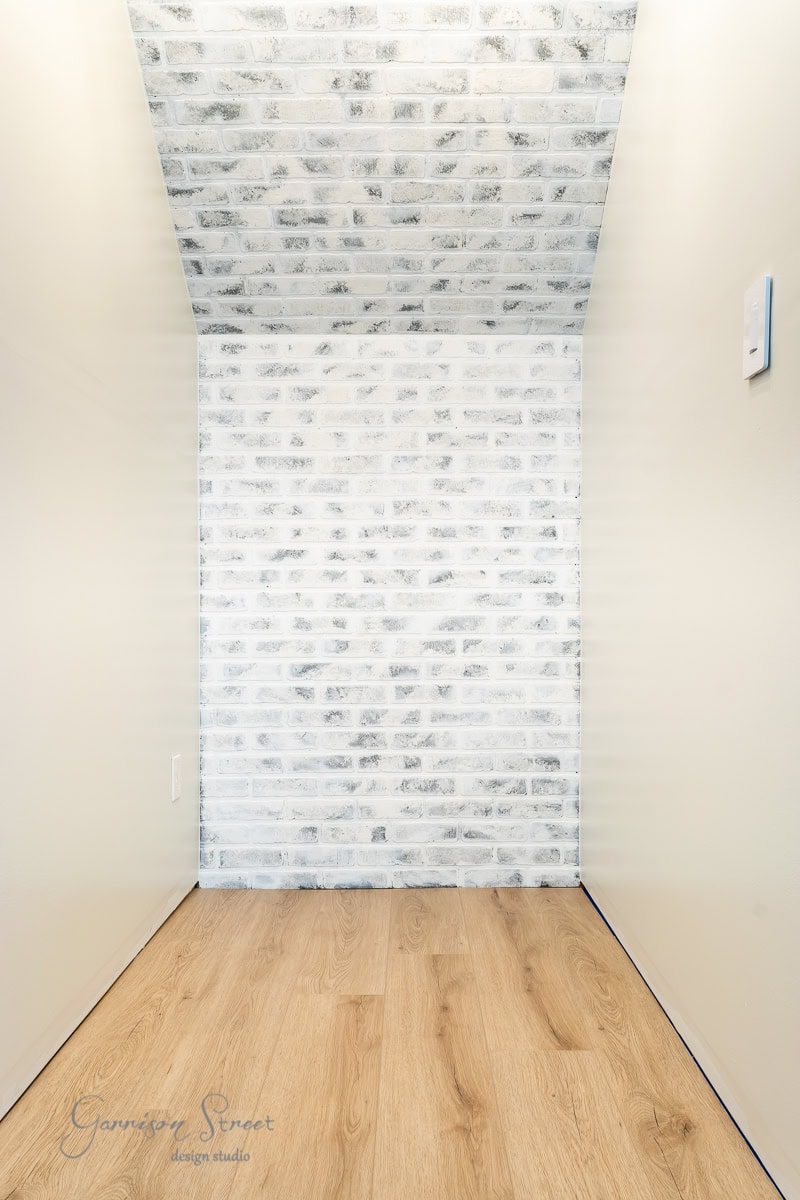 A minimalist hallway with light wood flooring and white brick walls features a small button on the right wall. Its narrow, well-lit space exudes a clean and modern vibe—perfect inspiration for those wondering how to build a DIY wine rack that complements such sleek design aesthetics.