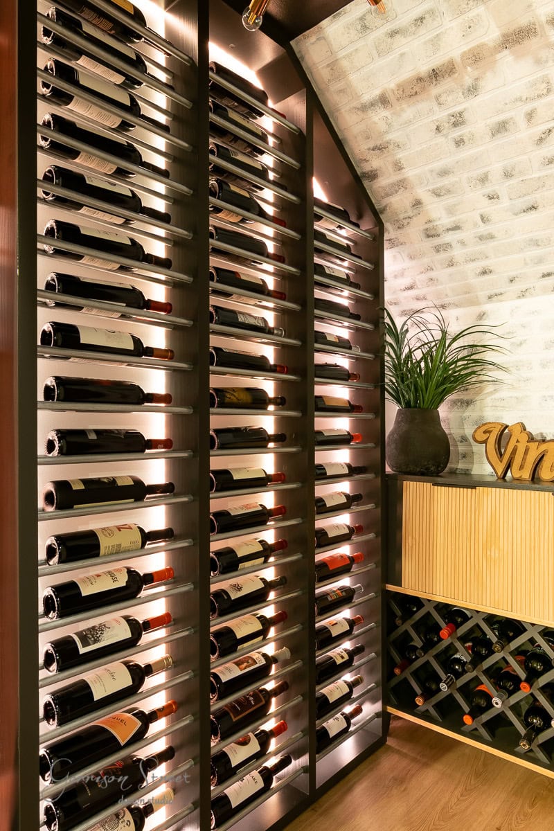 A stylish wine cellar with illuminated shelves displays numerous wine bottles. A plant in a dark pot sits near a custom-built DIY wine rack filled with more bottles. The room features a white brick wall and wooden flooring, creating a cozy atmosphere perfect for wine enthusiasts.