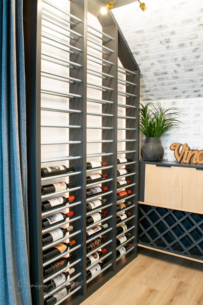 A wine storage area with a modern, vertical rack displays multiple bottles, showcasing how to build a DIY wine rack. The space features a light wood floor and textured white brick wall. A cabinet with a black lattice design and a potted plant adds an elegant touch.