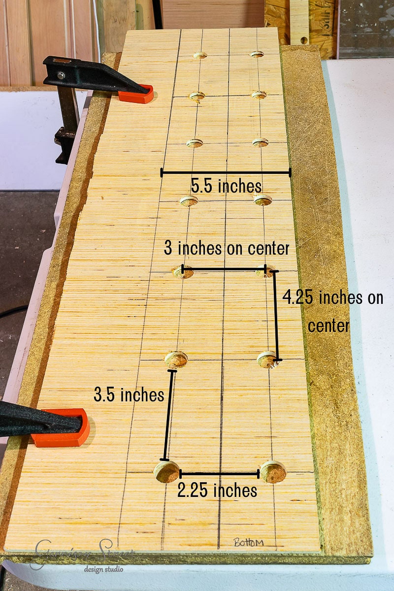 The wooden board clamped to the table, with drilled holes and precise measurements labeled (5.5 inches, 3 inches on center, 4.25 inches on center, 3.5 inches, and 2.25 inches), is a perfect starting point for learning how to build a DIY wine rack with ease and accuracy.