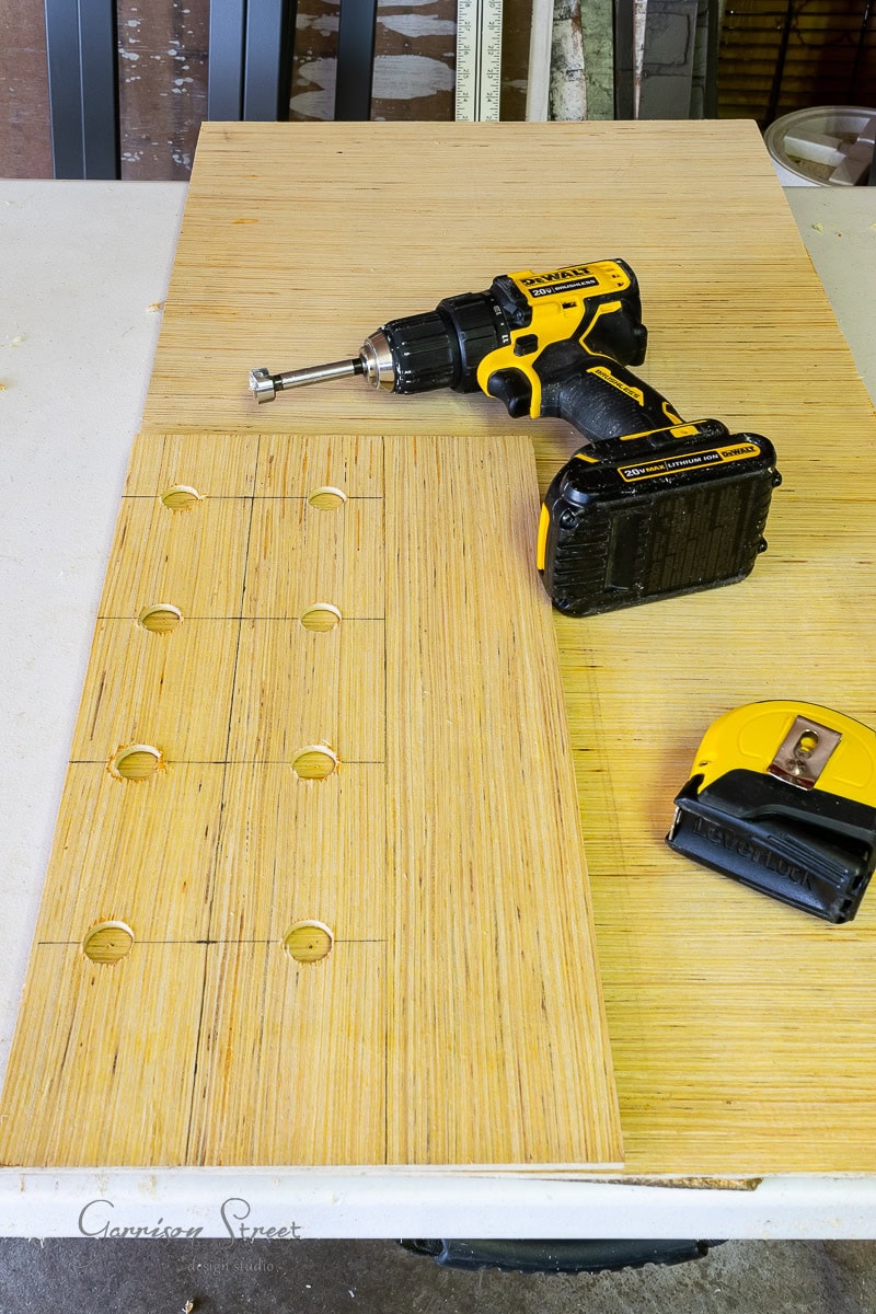 A workbench hosts a piece of plywood with multiple drilled holes, laying the groundwork for your DIY wine rack. A yellow and black cordless drill with a spade bit, alongside a trusty yellow tape measure, are poised and ready for use on this creative project.