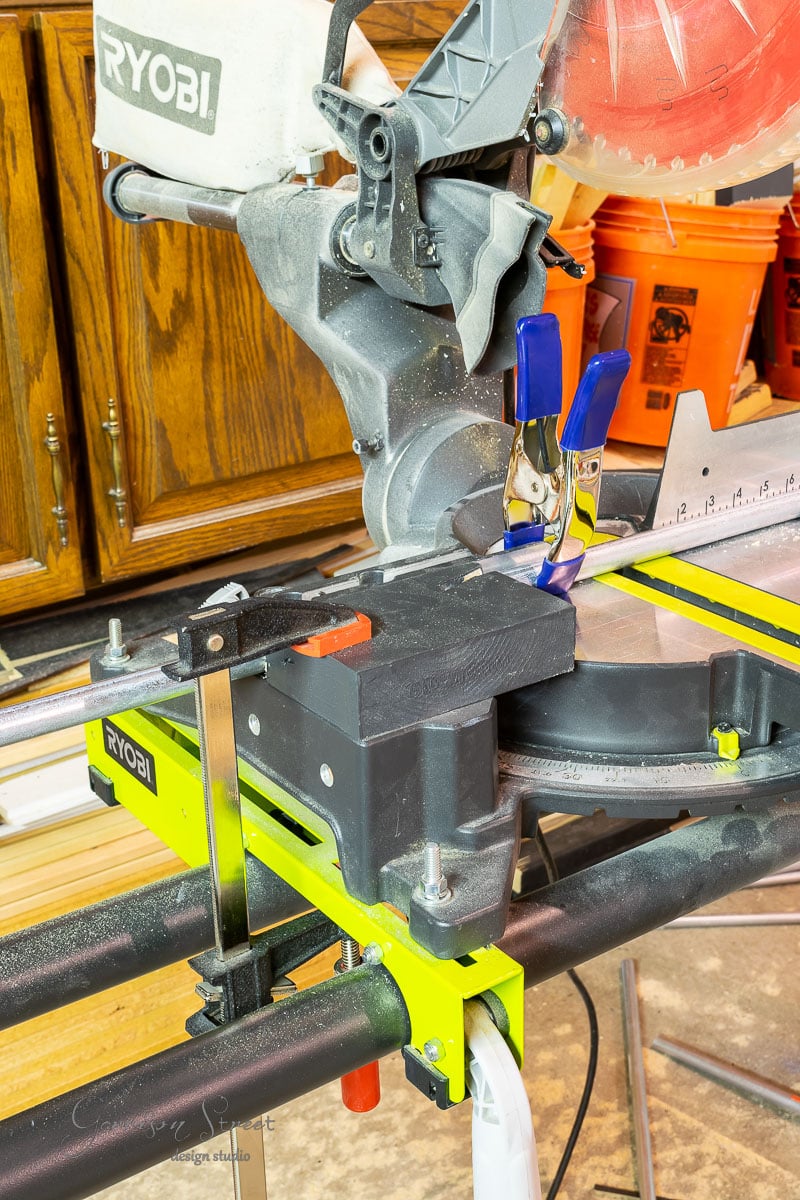 A Ryobi miter saw with a red blade is clamped onto a workbench, the perfect setup for your next project on how to build a DIY wine rack. Two blue clamps secure the material near the saw. In the background, wooden cabinets and a blurred orange bucket add to the workshop vibe.