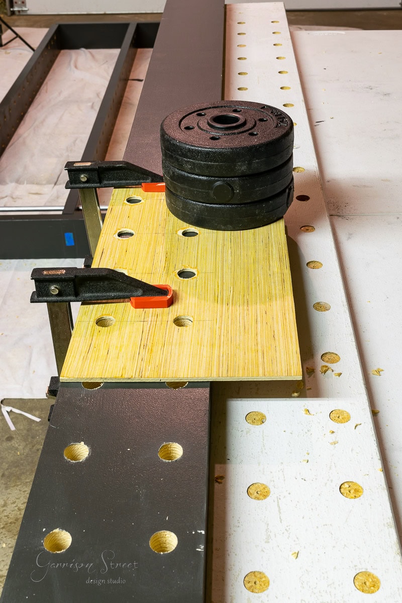 A piece of wood is clamped to a perforated metal workbench, likely part of a setup for how to build a DIY wine rack. Several weights are stacked on the wood, suggesting precision is key in this woodworking or construction project.