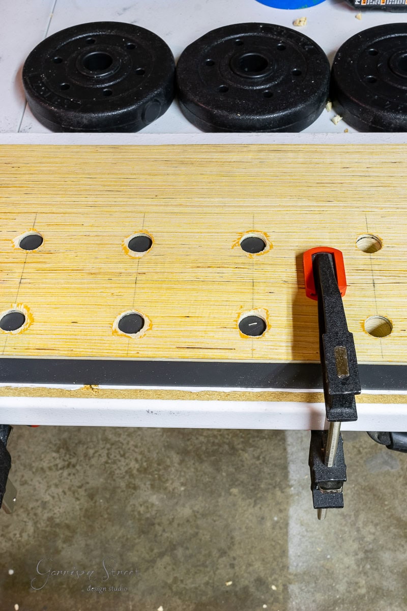 A wooden board with evenly drilled holes is clamped to a workbench, perfect for learning how to build a DIY wine rack. Three weight plates line the top edge of the table, resting on the sturdy concrete surface below.