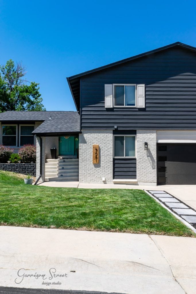 Home A contemporary house with dark gray siding and white stucco exterior. The entrance features a teal door with potted plants on either side. A paved driveway leads to a garage. The lawn is neatly manicured under a clear blue sky.