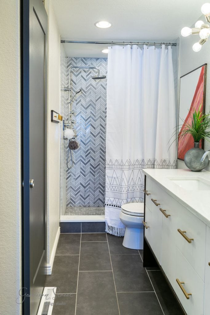 Home Modern bathroom with black tile flooring, a white vanity with gold handles, and a geometric-patterned shower curtain. The shower has a gray herringbone tile detail. A potted plant and decorative artwork are on the countertop.