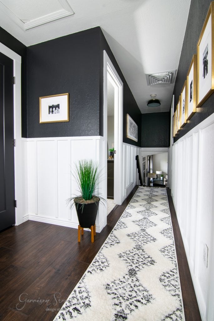 A modern hallway with dark walls and white wainscoting features framed pictures, a long patterned rug, and a tall plant in a black pot. The ceiling light illuminates the narrow space leading to a room.