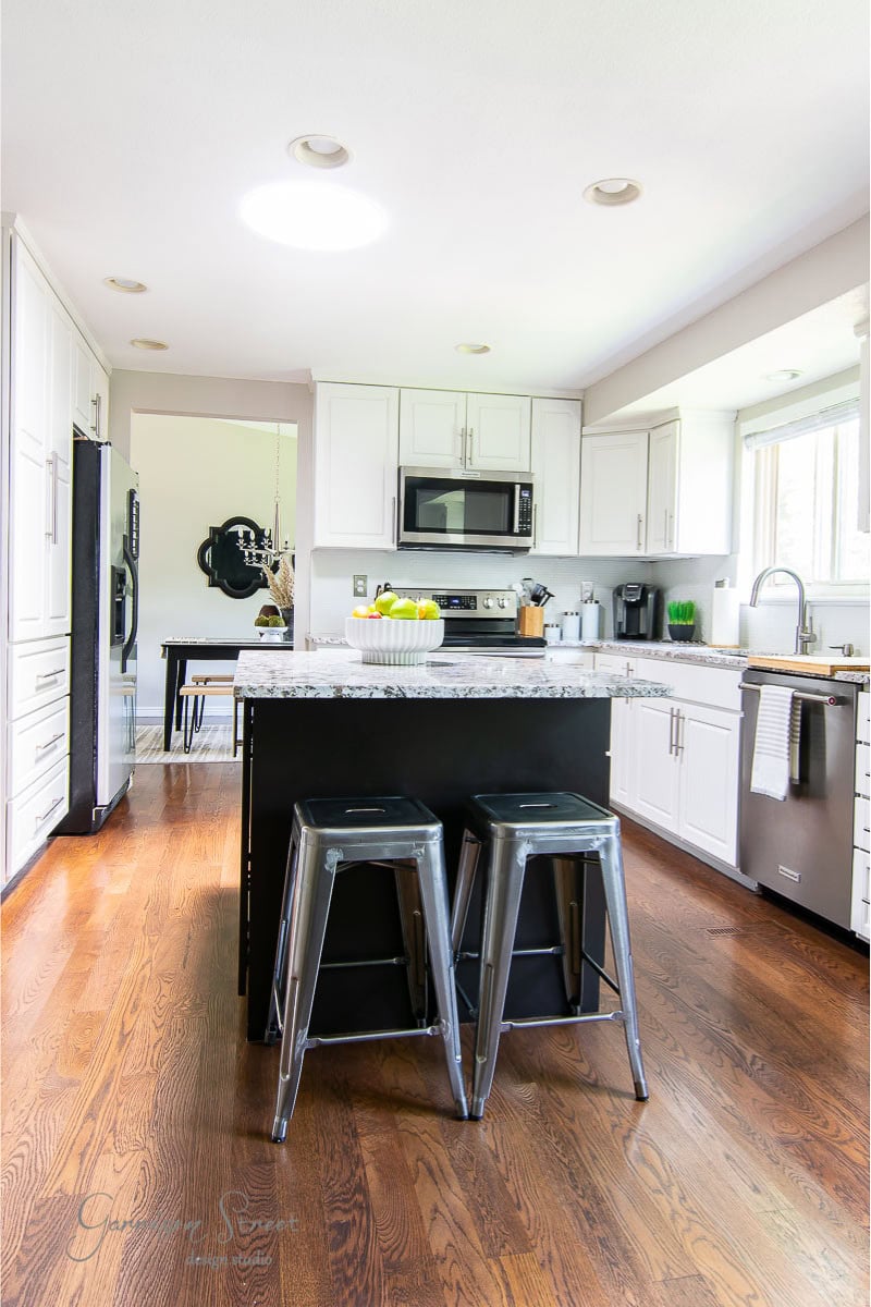 Modern kitchen with white cabinets, marble countertop island, and two metal stools. Stainless steel appliances, including a microwave and dishwasher, are present. The space has large windows offering natural light and hardwood floors throughout.