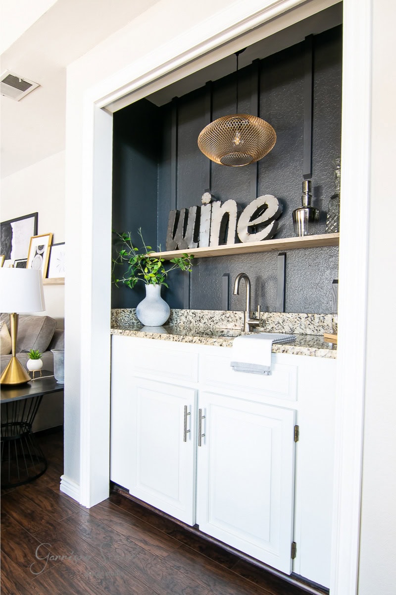 A small home bar with white cabinets, granite countertop, and a modern black backsplash. A metal "wine" sign and bottles are showcased on a wooden shelf. A stylish pendant light hangs above, while a large gray vase with greenery adorns the counter.