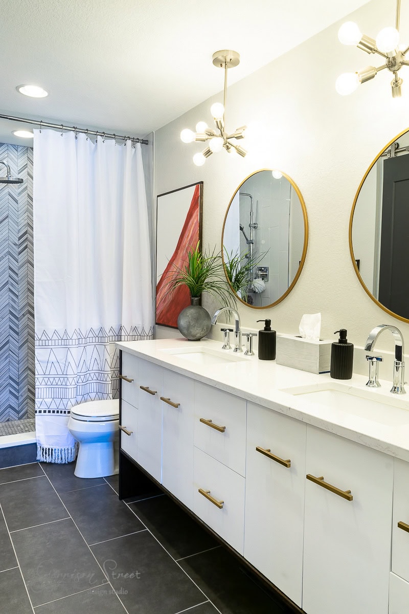 Modern bathroom with dual sinks and round mirrors framed in gold. A large white vanity boasts black and gold fixtures, while a shower with a chevron-patterned curtain stands in the background. A potted plant rests on the counter.