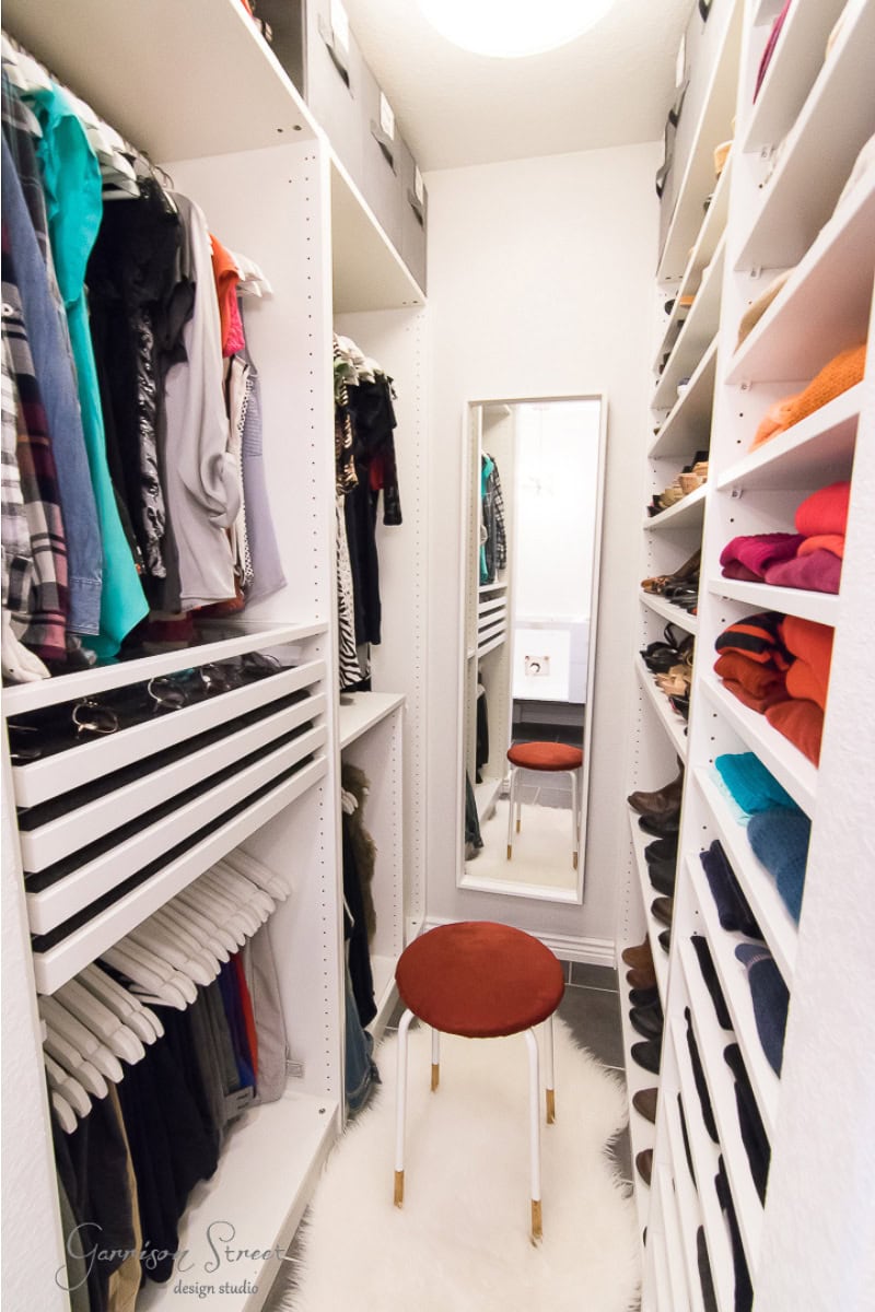 A walk-in closet with shelves filled with neatly arranged clothes and shoes. A narrow aisle leads to a full-length mirror at the back, while a round stool with a red cushion sits elegantly on a white rug in the center.