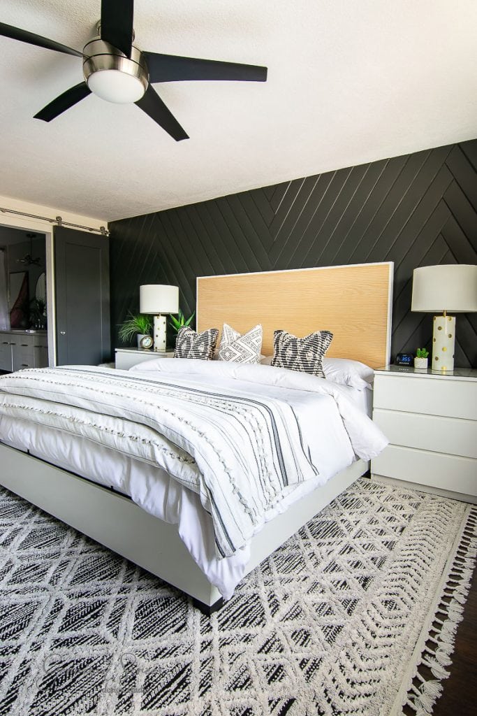 Modern bedroom with a large bed featuring white and striped bedding—a great inspiration for remodeling ideas for split level houses. The dark back wall has a chevron pattern, and a ceiling fan hangs above. Two white nightstands with lamps flank the bed, while a patterned rug covers the floor.