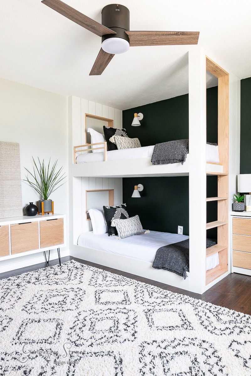 Modern bedroom with bunk beds built into a white wall, featuring dark green accents and wooden shelves. A light fixture is overhead, and the room is decorated with a lush rug, plants, and contemporary furniture.