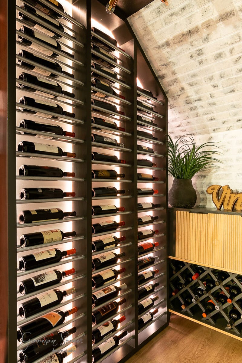A wine cellar with a vertical rack brims with bottles. Wooden floors meet brick walls, while a small plant and a "Vino" sign add charm on the right.
