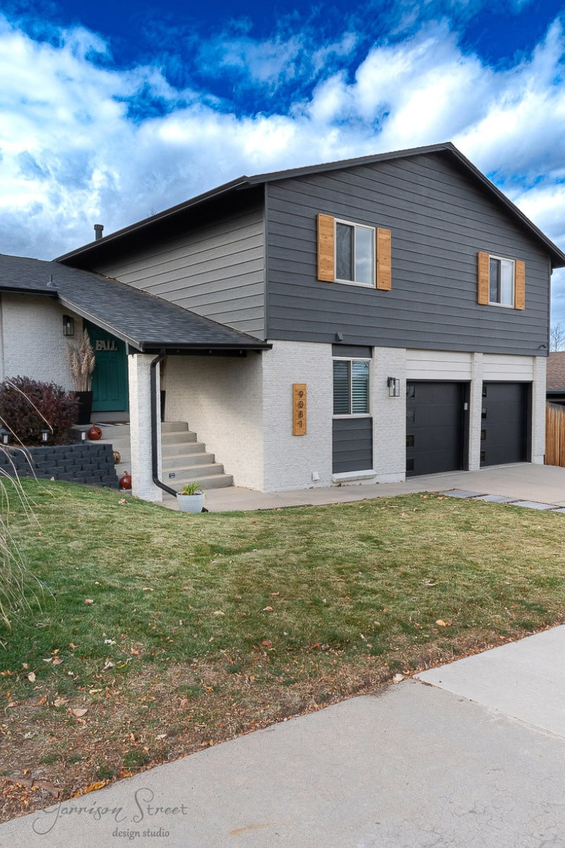 A modern two-story house with a gray and white exterior, wooden shutters, and a double garage. A well-kept lawn graces the foreground with a pathway leading to the entrance under a partly cloudy sky.