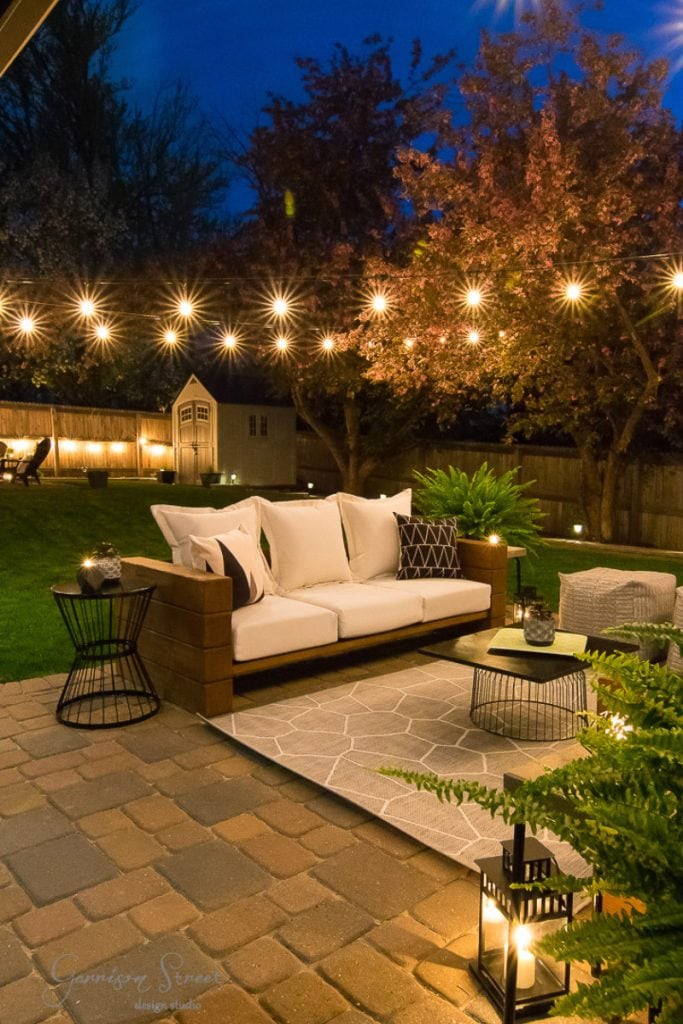 A cozy backyard patio at dusk features a wooden sofa, white cushions, and black side tables on a brick floor. String lights hang overhead, suggesting remodeling ideas for split-level houses. A small shed nestles in the background amid trees and a grassy area.