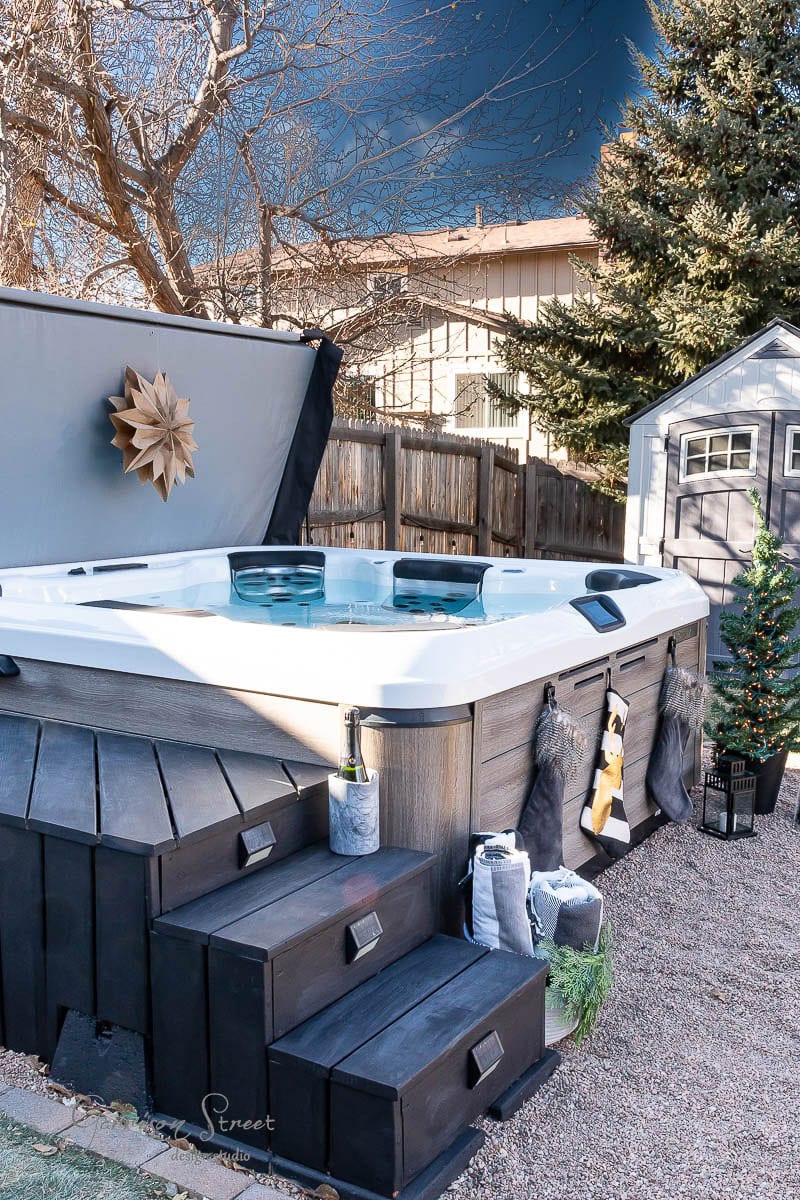 A backyard oasis offers a filled hot tub with open lid, encircled by wooden steps perfect for relaxation. Towels and a champagne bottle invite indulgence, while a small decorated tree adds charm. Beyond, a shed and trees complete the scene.