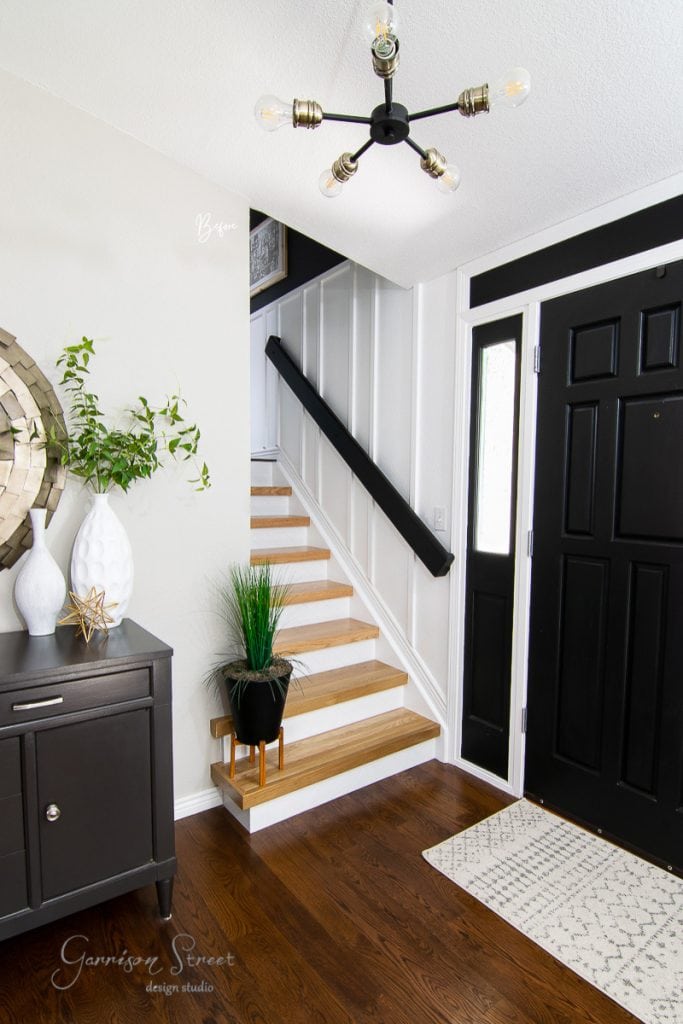 Modern entryway with a black door, wood flooring, and white walls—perfect inspiration for remodeling ideas for split-level houses. A staircase with wood steps and a black railing enhances the space, as does a sleek sideboard adorned with decorative vases and plants. A contemporary light fixture hangs overhead.