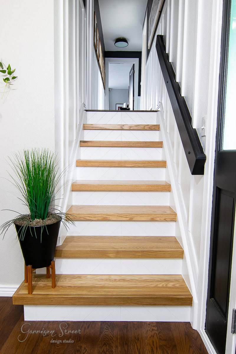 A modern staircase with wooden steps and white risers, flanked by sleek white railings. A black handrail adds contrast on the right side, while a potted green plant at the base enhances the welcoming vibe.