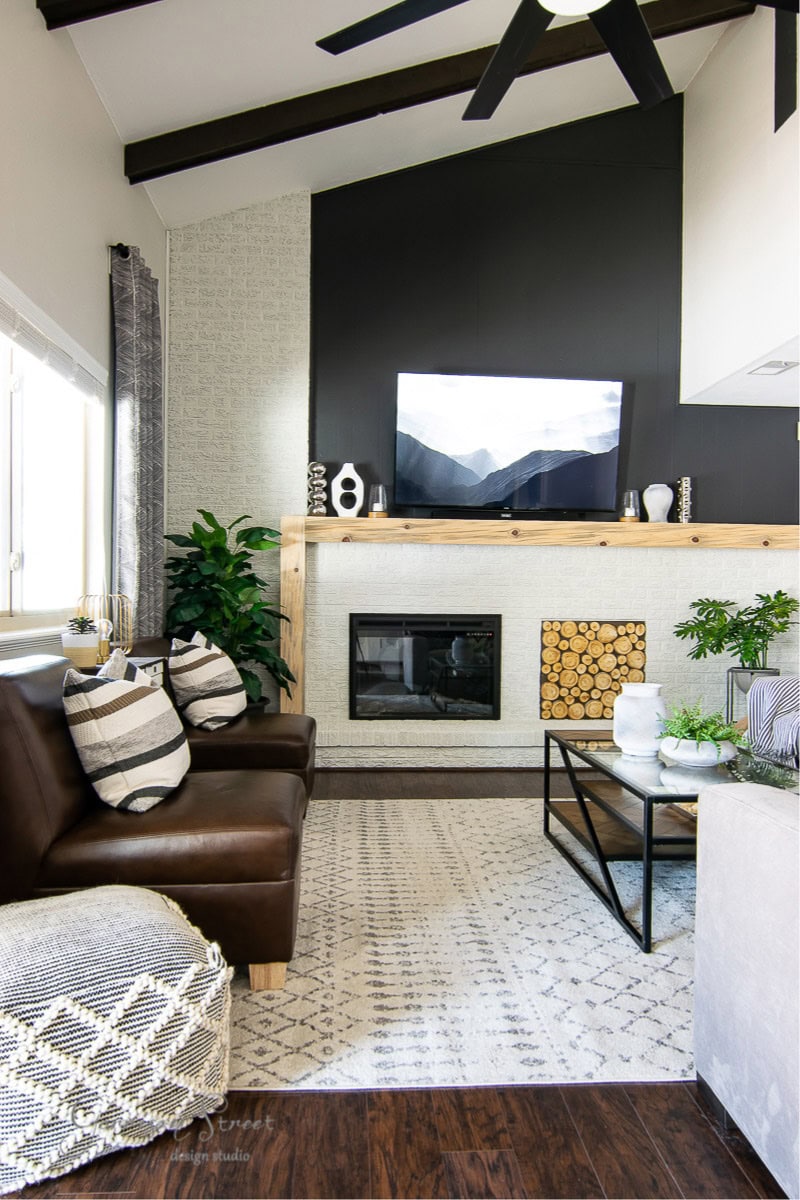 A modern living room with a dark accent wall featuring a mounted TV and fireplace. The space has a brown leather sofa with striped pillows, a patterned rug, wooden coffee table, decorative plants, and logs stacked by the hearth.