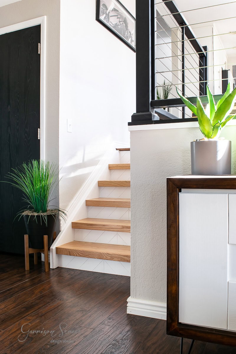 Modern interior with wooden stairs. A sleek white sideboard with a potted plant sits in the foreground. Bright light fills the room, highlighting the dark wooden floors and contrasting with the white walls and railing.