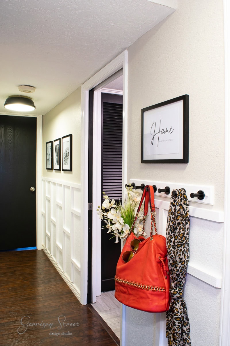 The hallway features white wainscoting, dark wood flooring, and black doors. Wall art includes a "Home" sign and monochrome photos. A red bag and leopard print scarf hang on hooks, while a bouquet of white flowers decorates a side table.