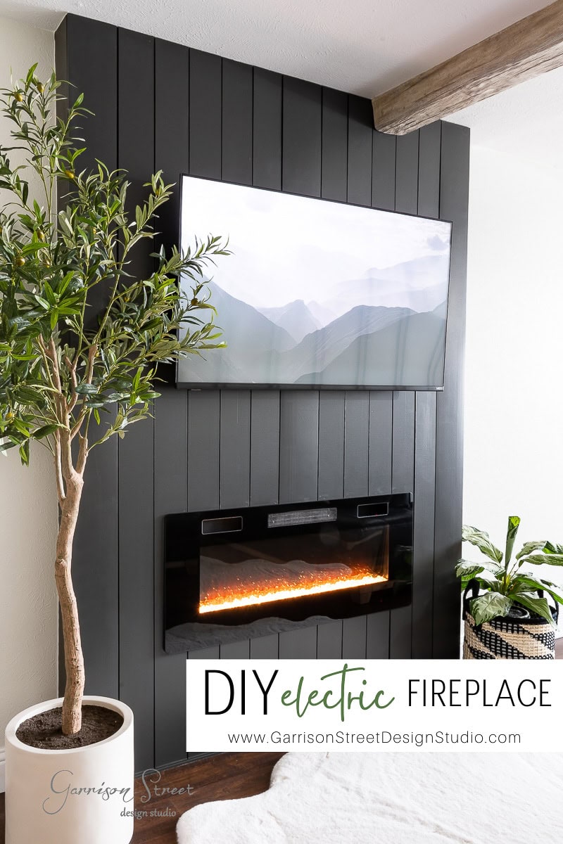 A modern electric fireplace and TV wall is featured with a flat-screen mounted above on a dark vertical shiplap backdrop. Potted plants flank the setup, infusing greenery into the sleek, minimalist decor. The text reads "DIY electric FIREPLACE" with a website link below.