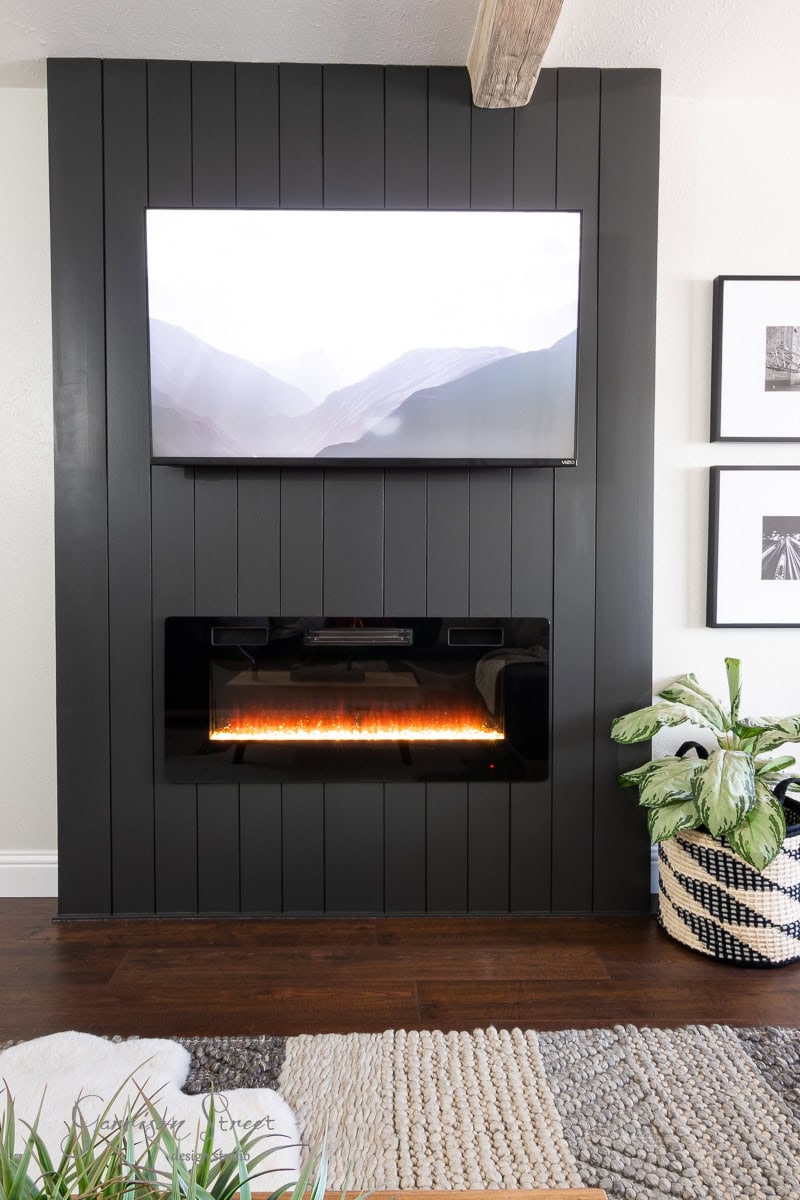 A modern living room boasts a sleek black paneled wall, showcasing a DIY electric fireplace and TV wall. The mounted flat-screen TV sits above the contemporary fireplace, with a potted plant and textured basket to the side, all complemented by a cozy rug on the wooden floor.