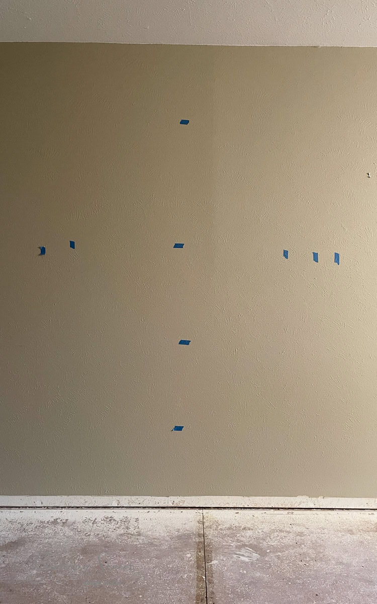 A beige wall with several pieces of blue painter's tape arranged in a cross pattern, serving as a guide for a DIY electric fireplace and TV wall. The floor below has a rough, unfinished texture.
