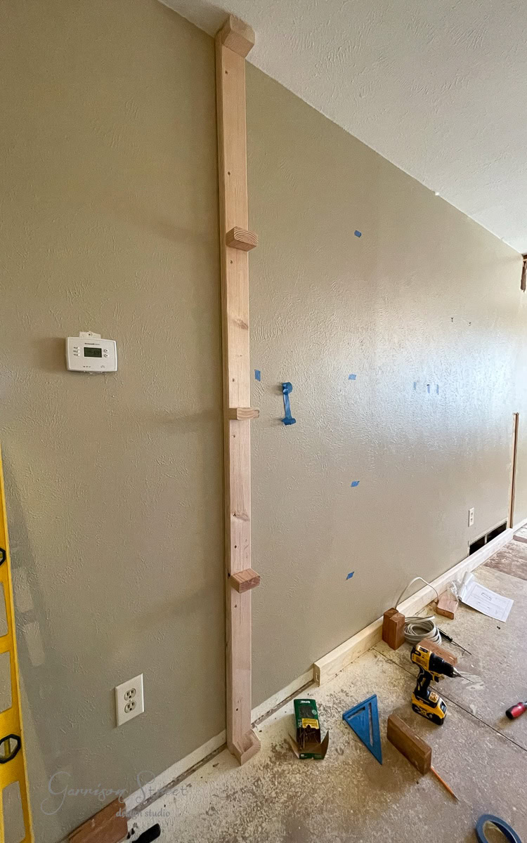 A partially constructed floating shelf being installed on a beige wall hints at a future diy electric fireplace and TV wall. Wooden brackets are attached vertically, with tools like a drill and measuring tape scattered below. An electrical outlet and thermostat are visible nearby.