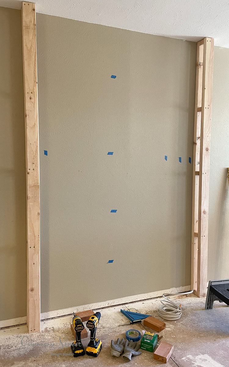 A partially constructed wall with vertical wooden beams on each side hints at a future DIY electric fireplace and TV wall. Unpainted drywall, marked with small blue tape pieces, fills the center. Tools, including a drill, gloves, and measuring instruments, rest on the floor.
