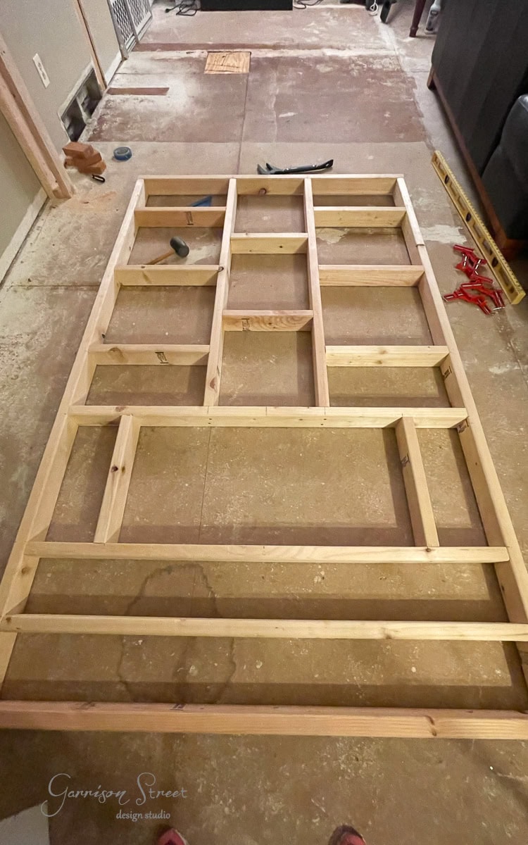 A wooden grid-like frame is laid on a bare floor, surrounded by various tools like a level, hammer, and pliers. The room appears under renovation, with concrete flooring and scattered materials hinting at the creation of a DIY electric fireplace and TV wall.