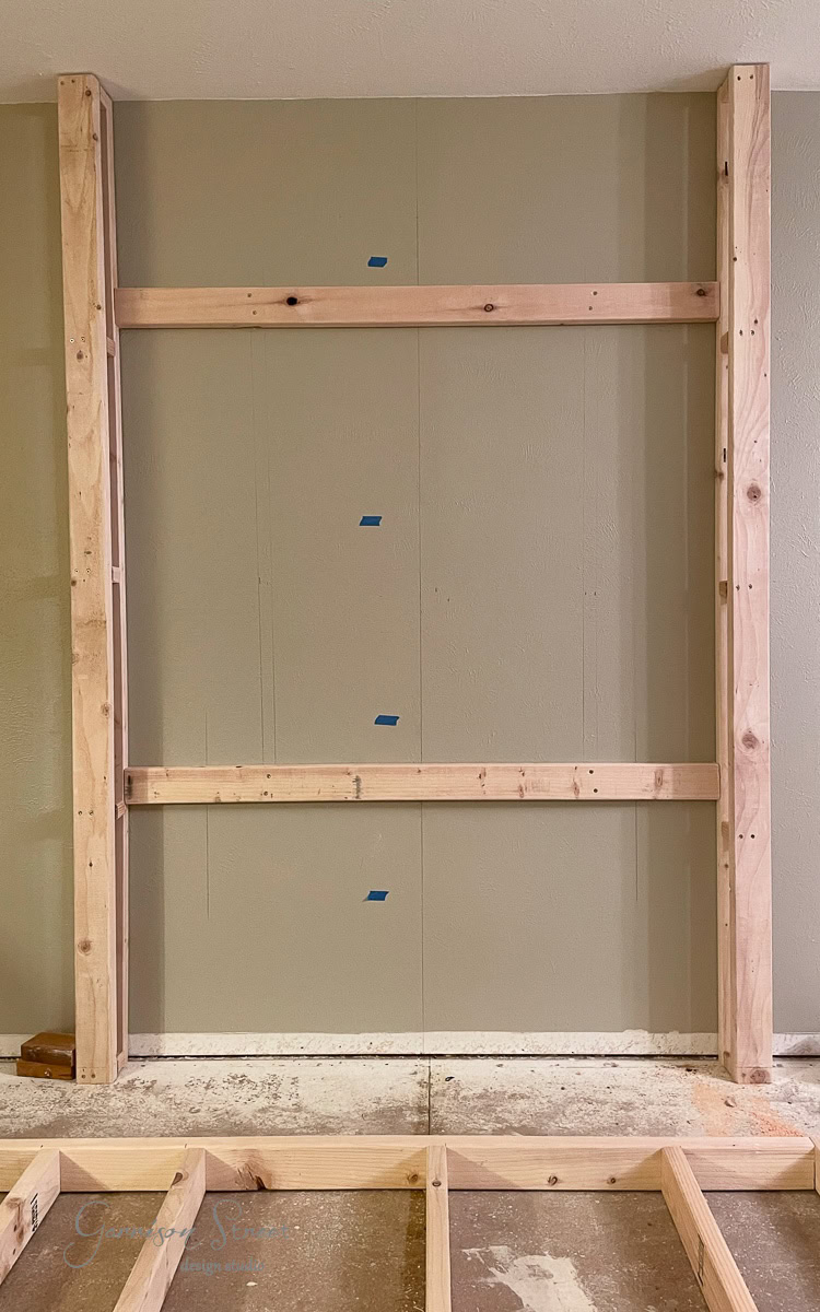 A wooden framing structure is being crafted for a DIY electric fireplace and TV wall. The floor showcases visible marks and measurements, while small blue tape pieces line the wall, indicating this is part of an intricate construction or renovation project.