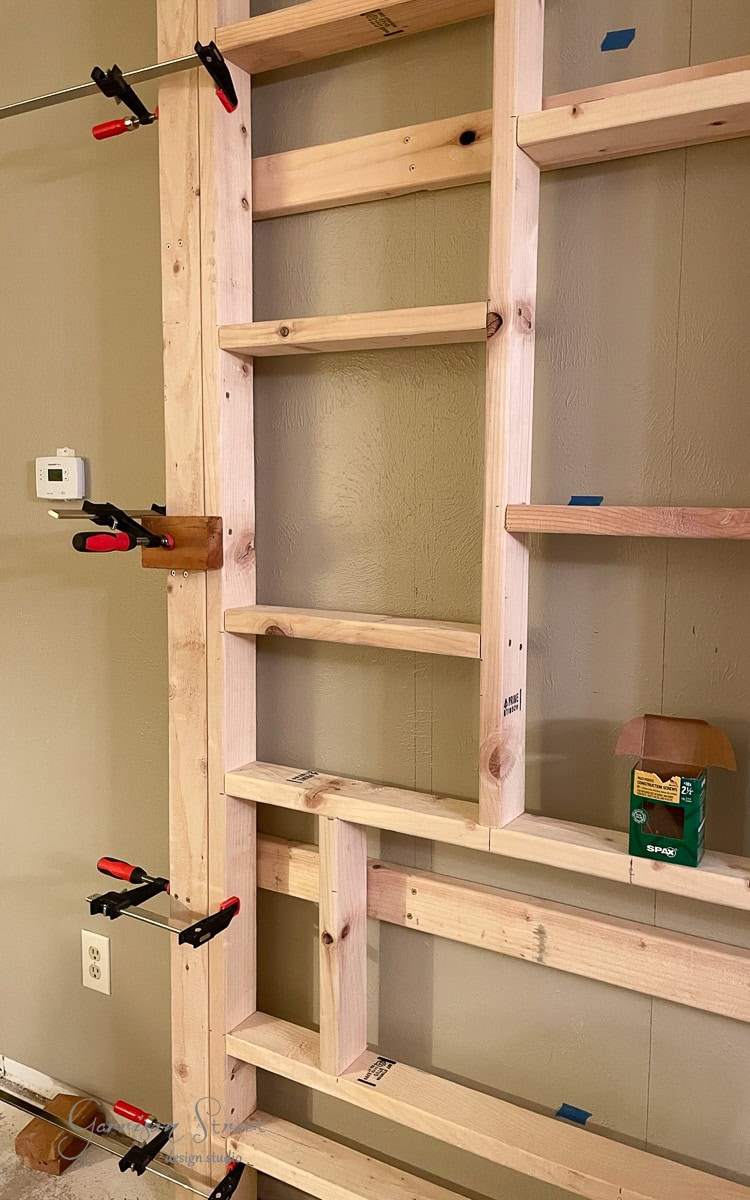 A wooden frame structure is attached to the wall, holding pieces in place with several clamps. Nestled beside a DIY electric fireplace and TV wall setup, a box of screws sits on a ledge. A light switch and outlet are visible nearby, ready for installation.