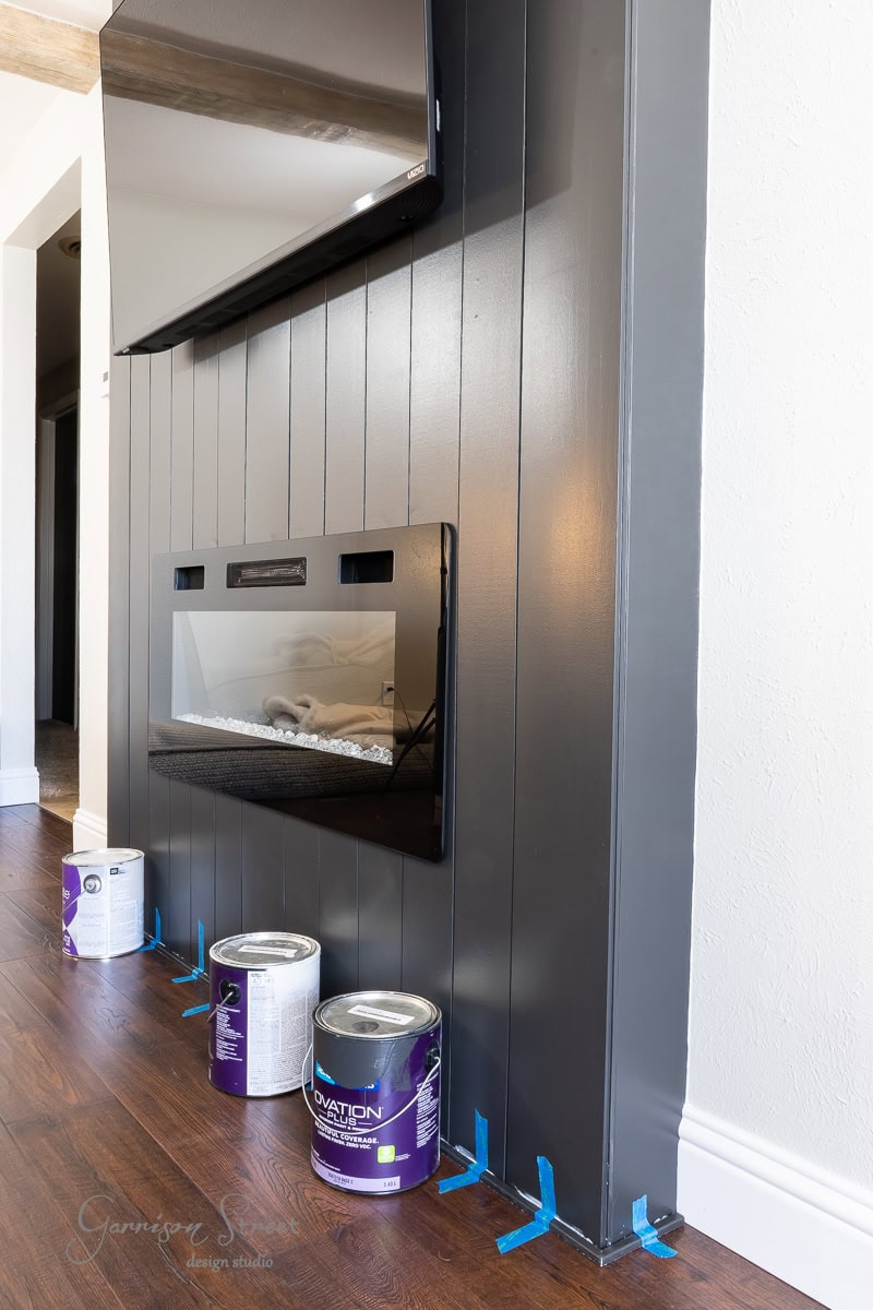 A modern interior with a sleek DIY electric fireplace set in a vertical panel wall. Three paint cans and painter’s tape rest on the dark hardwood floor, hinting at ongoing renovations. A TV is mounted above the fireplace, creating a stylish entertainment wall.