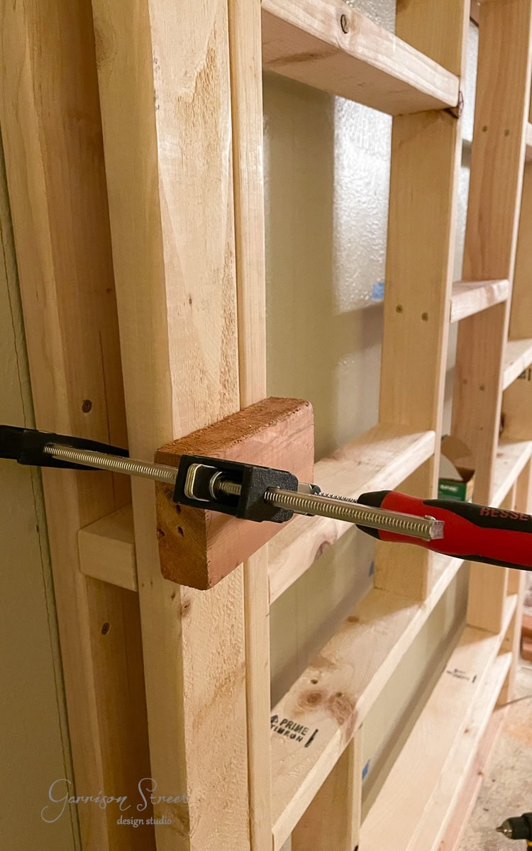 A wooden shelf frame under construction is shown with a clamp securing a horizontal block. The unfinished wood and tools suggest an ongoing DIY project, possibly part of crafting a custom electric fireplace and TV wall unit.