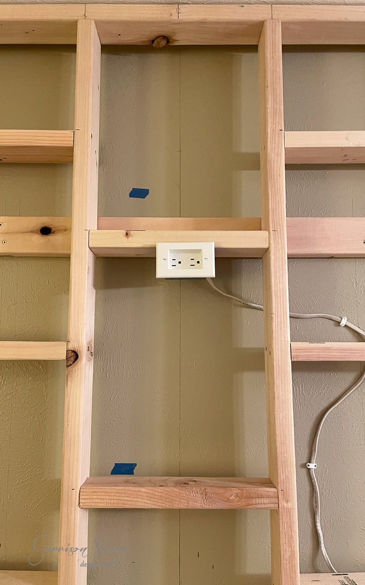 A wooden framework on a beige wall showcases unpainted shelves and a central outlet with plugged-in white cables, hinting at a DIY electric fireplace and TV wall project. Blue tape marks are visible on the wood, adding to the work-in-progress charm.