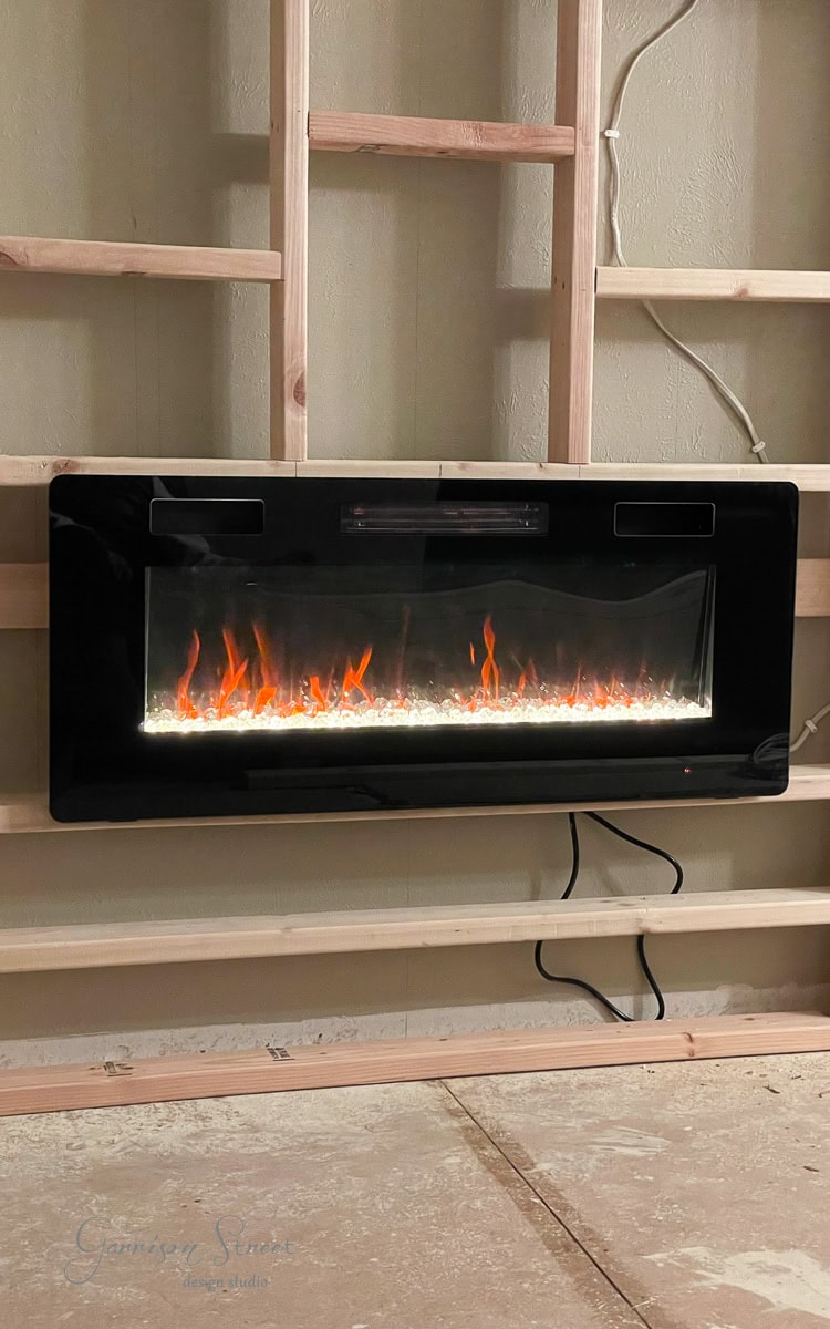 A modern electric fireplace with a sleek black frame is installed within a wooden shelf structure, perfect for a DIY electric fireplace and TV wall project. Orange flames and glowing embers flicker through the glass front, set against an unfinished backdrop of visible wires and wooden beams.