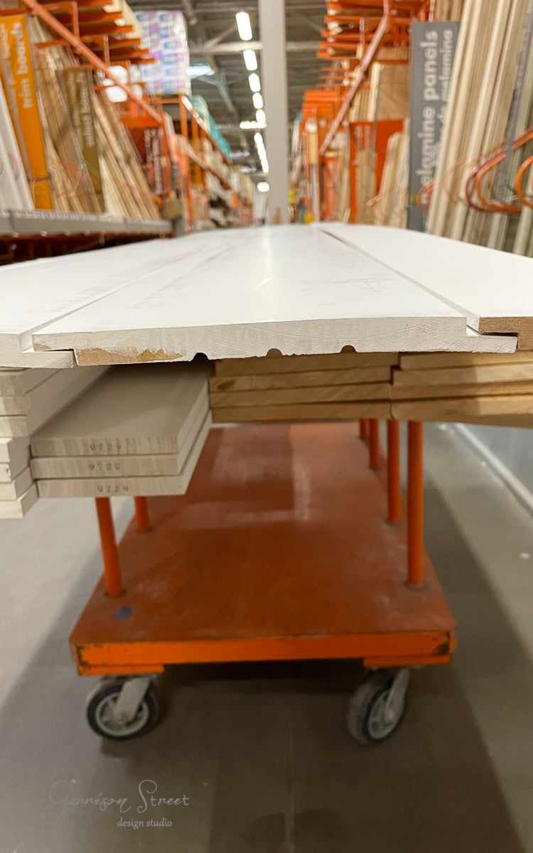 A stack of white wooden boards rests on an orange wheeled cart in a hardware store aisle, perfect for crafting your DIY electric fireplace and TV wall. The cart glides across the polished concrete floor, flanked by shelves filled with various building materials.