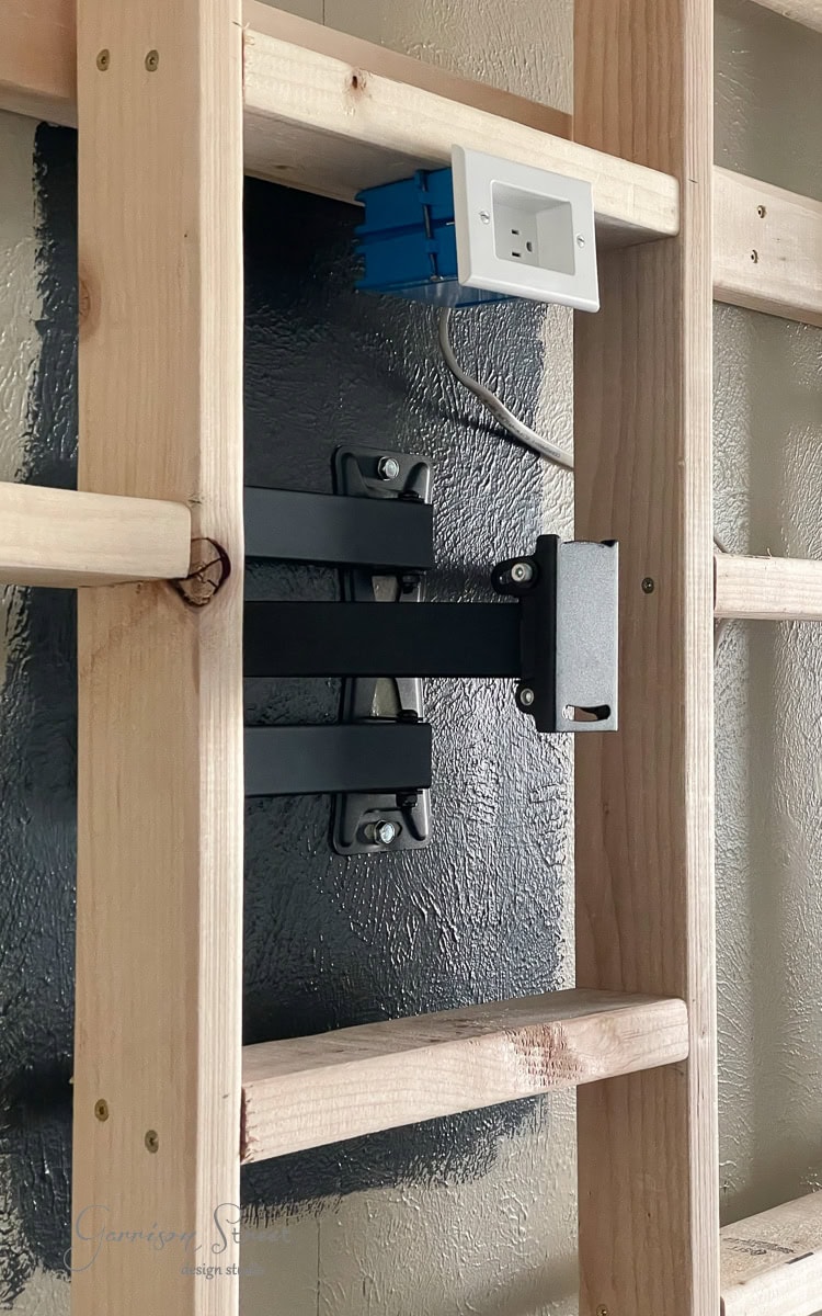 Wooden wall framework with a metal bracket attached, ideal for mounting a TV or monitor on your DIY electric fireplace and TV wall. An open blue electrical box with a white outlet is mounted on a horizontal piece of wood, ready to power your setup.